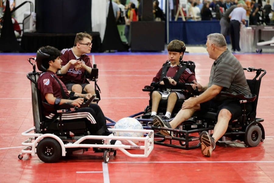 Coach Brian Broyles with Rolling Rapids athletes in wheelchairs gathered around a power soccer ball during a Nu-Venture clinic